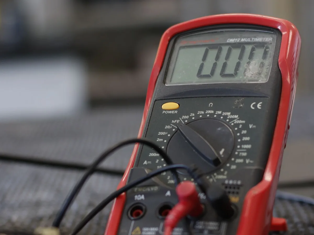 Ein digitales Multimeter mit angeschlossenen Messleitungen steht auf einer Arbeitsunterlage, im Hintergrund unscharfe Bauteile.