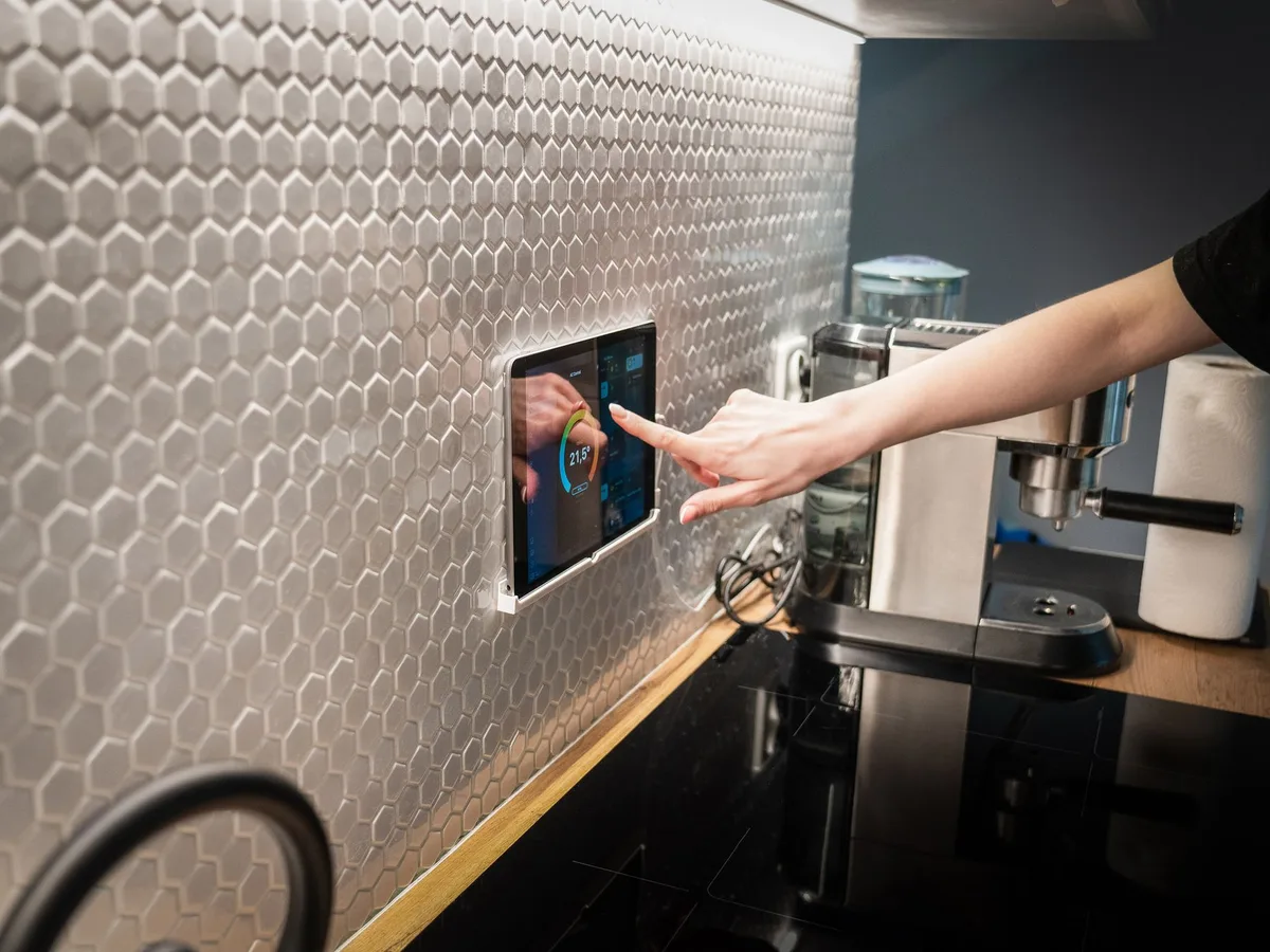 Eine Hand bedient ein wandmontiertes Smart-Home-Bedienpanel mit Touchscreen, das die Raumtemperatur anzeigt.