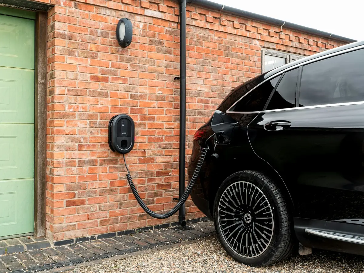 Ein schwarzes Elektroauto lädt an einer wandmontierten Ladestation an einer Backsteinfassade, das gewendelte Ladekabel führt zum Fahrzeug.
